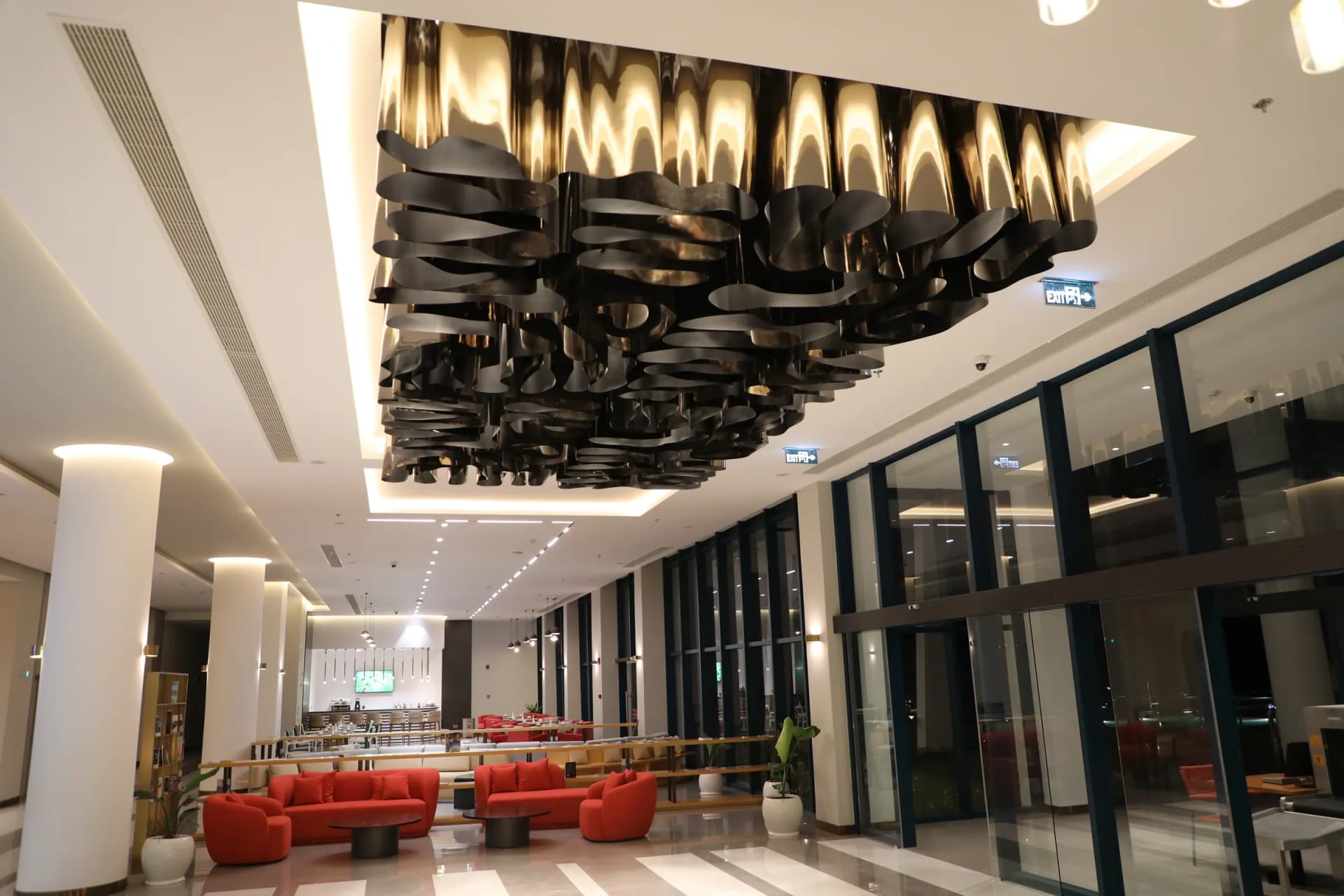 Lobby bar lounge crowned by a sculptural bronze-ribbon ceiling installation above coral velvet sofas and marble-striped floors